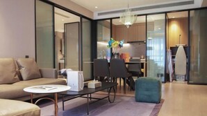 Una sala moderna con sofá, mesa de centro y reposapiés, un área de comedor con sillas, una cocina al fondo y un ventanal grande.