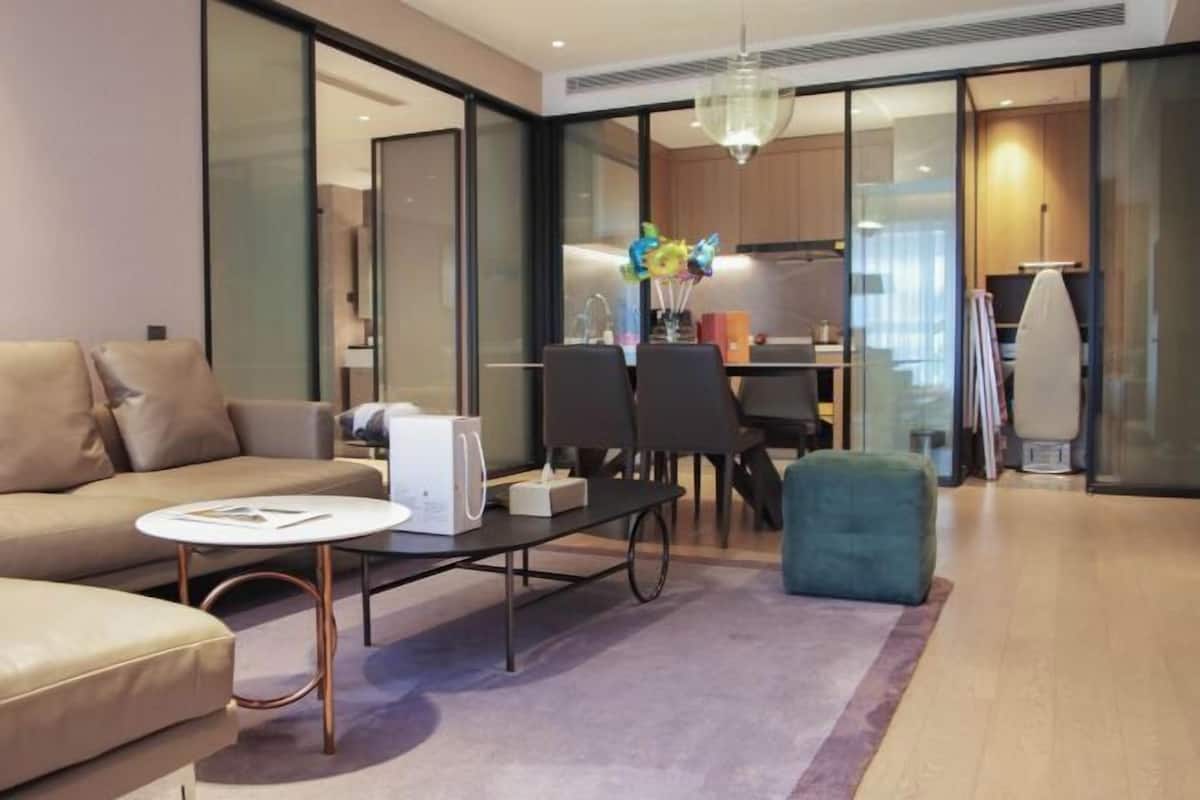 A modern living room with a sofa, coffee table, and ottoman, a dining area with chairs, a kitchen in the background, and a large window.