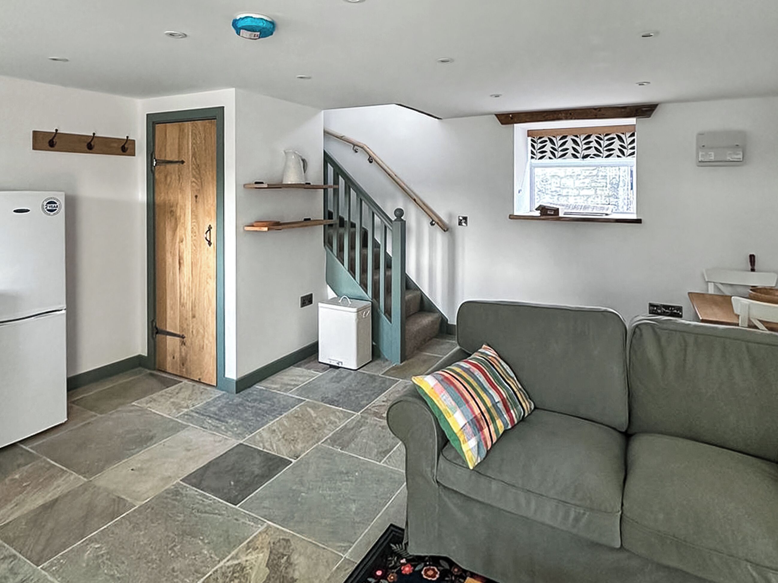 A compact living space with a kitchenette, a grey sofa with a striped pillow, a wooden door, a staircase, and a window with a decorative screen.