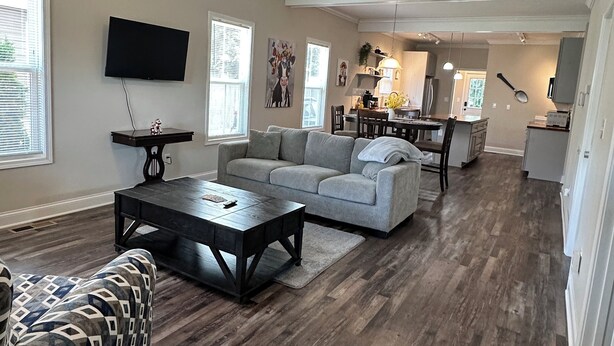 A spacious living room with a grey sofa, a patterned armchair, and a wooden coffee table. A ceiling fan and a TV are mounted on the wall.