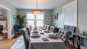 Een eetzaal met een lange tafel gedekt voor een maaltijd, een kroonluchter en een groot raam met jaloezieën.