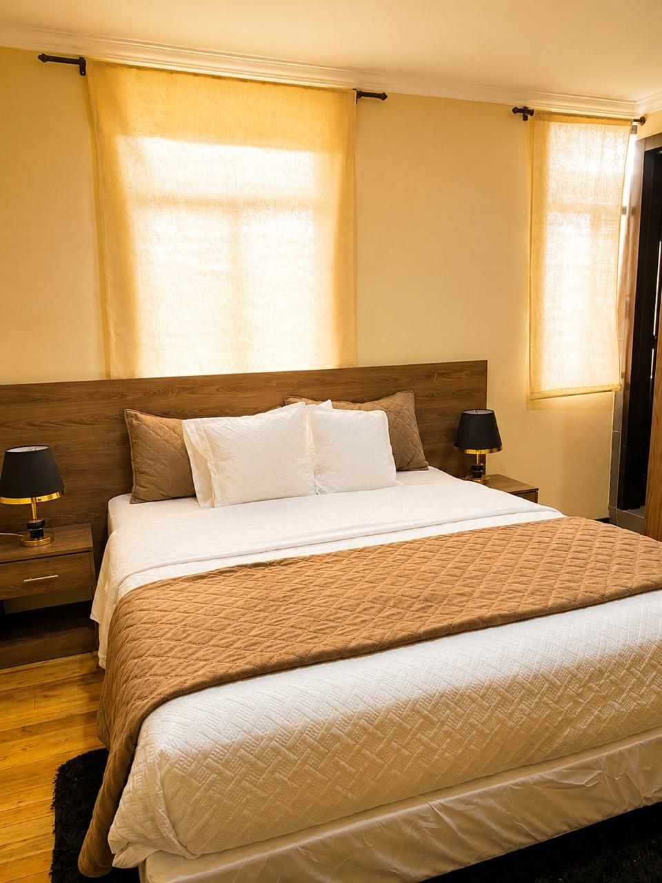 A hotel room with a large bed, two bedside tables with lamps, a wooden headboard, and curtains on the windows.