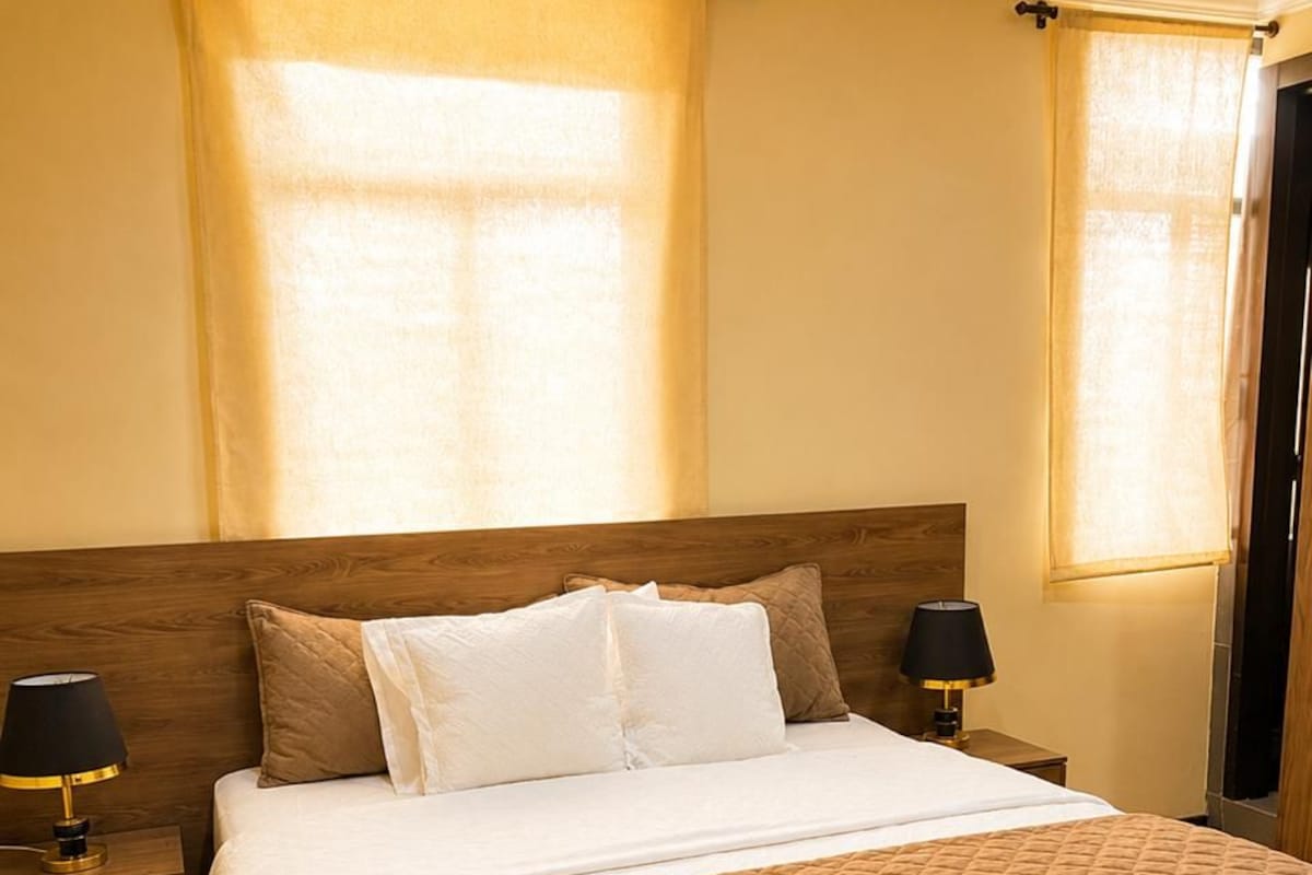 A hotel room with a large bed, two bedside tables with lamps, a wooden headboard, and curtains on the windows.
