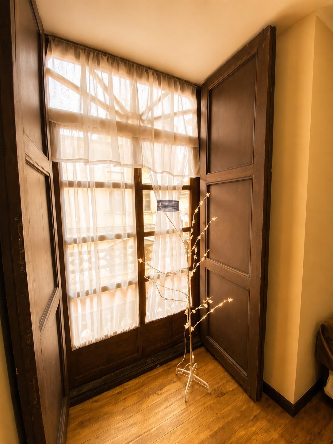 A room with a wooden floor, a window with sheer curtains, and a decorative light stand.