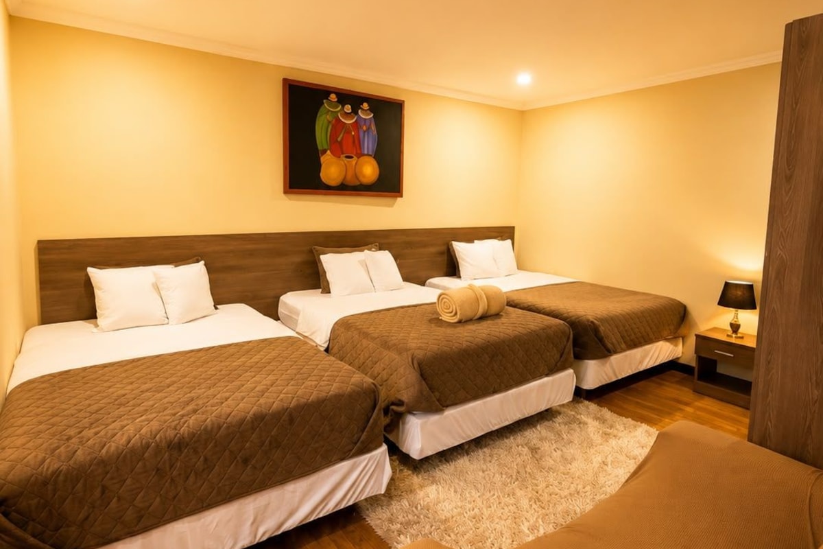 A hotel room with three beds, a wooden floor, a painting on the wall, and a nightstand with a lamp.