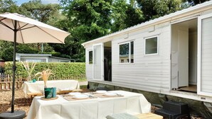 A mobile home with a dining area set up outside, featuring a table with plates and a tablecloth, a beige umbrella, and a bench. - Gorgeous home in Merksplas (Merksplas)