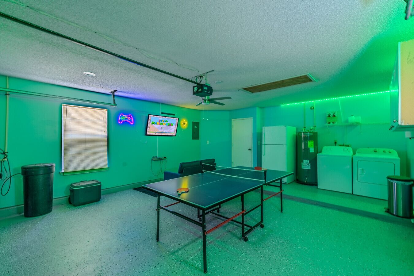 A room with a ping pong table, a ceiling fan, a TV, neon signs, and a washing machine.
