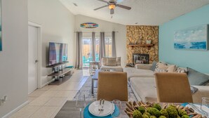 A spacious living room with a flat-screen TV, a stone fireplace, and a dining area set for a meal. - Beach Retreat with Backyard Flair! w/Yard Games (Atlantic Beach)