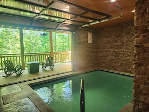 An indoor swimming pool with a wooden deck, two rocking chairs, and a stone wall.
