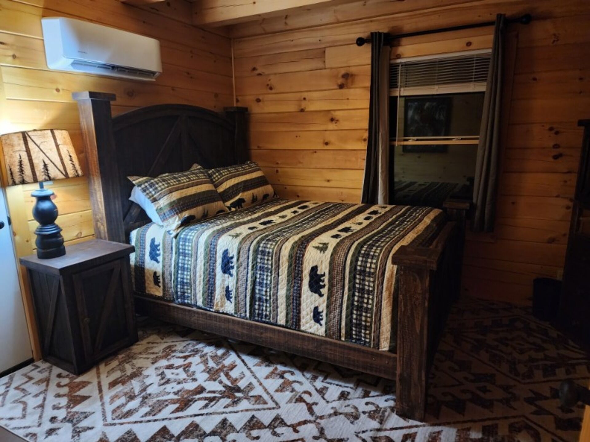 A wooden cabin bedroom with a bed, a nightstand, a lamp, and a window with curtains.