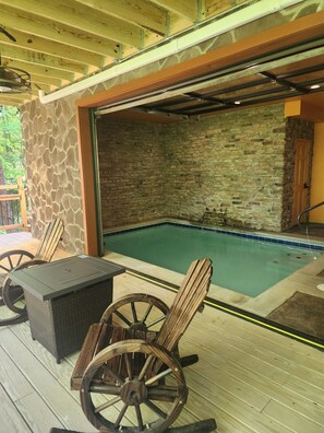 An outdoor pool area with stone walls, a wooden deck, and a table with chairs. - Brand new 2024 3bedroom indoor Pool cabin 1 acre private wooded lot Gatlinburg. (Gatlinburg)