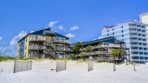 A beachfront hotel with multiple balconies, a sandy beach in the foreground, and a clear blue sky. - Sandpiper 7-C  ON THE BEACH - Heated Pool - Smart TVs (Gulf Shores)