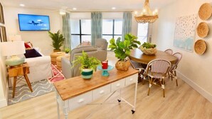 A modern living room with a sofa, a dining table with chairs, a TV mounted on the wall, and a chandelier. - Sandpiper 7-C  ON THE BEACH - Heated Pool - Smart TVs (Gulf Shores)
