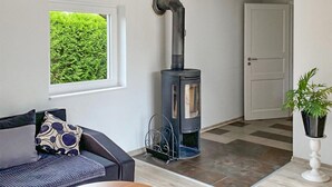 Un salon chaleureux avec un canapé, une petite table, un poêle à bois noir, une fenêtre donnant sur la verdure et une porte blanche.
