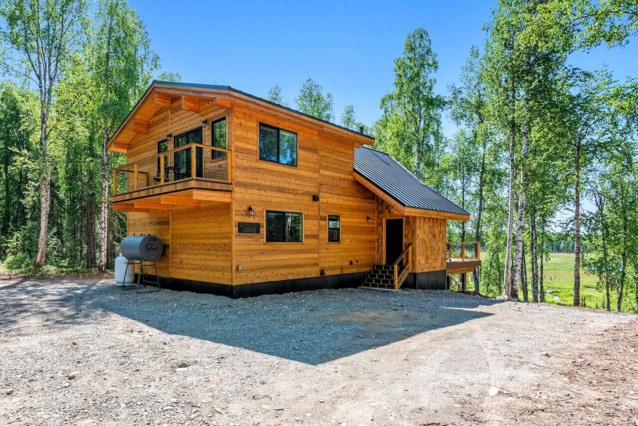 A two-story log cabin with a balcony, a covered porch, and a gravel driveway.