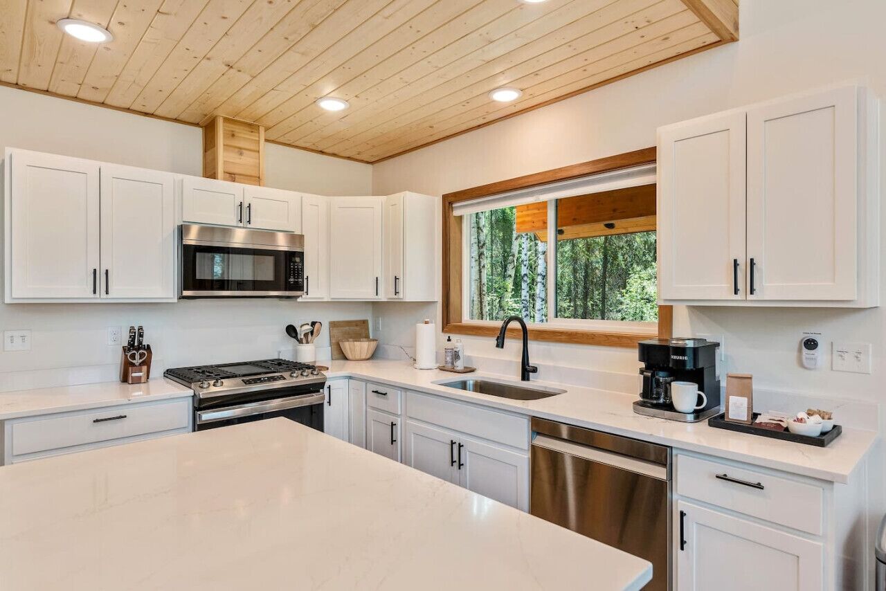 Una cocina moderna con armarios blancos, electrodomésticos de acero inoxidable y un amplio ventanal con vista a árboles.