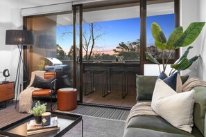 A modern living room with a sofa, a coffee table, and a floor lamp. There is a sliding glass door leading to an outdoor area with a barbecue setup.