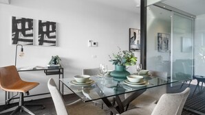 Een moderne eethoek met een glazen tafel, witte stoelen en een blauwgroene vaas met planten.