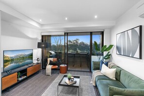 A modern living room with a flat-screen TV, a coffee table, a sofa, and a balcony with a view of the mountains. - Luxurious Fitzroy Street Getaway (St Kilda)