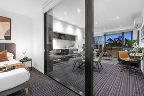 A modern hotel room with a bed, a dining area, and a kitchen visible through a glass partition.