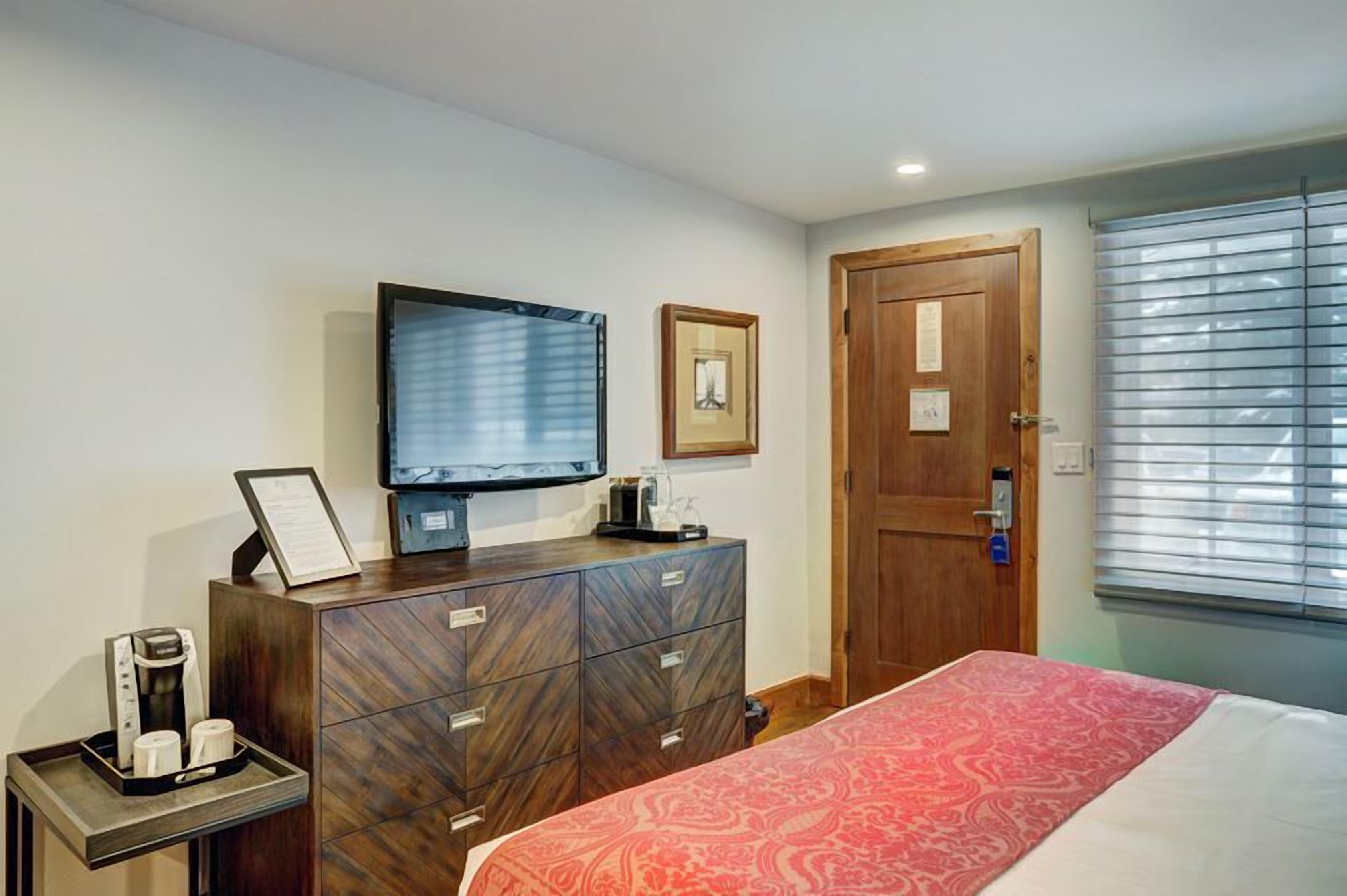 A hotel room with a bed, a TV, a wooden dresser, a coffee maker, and a window with blinds.