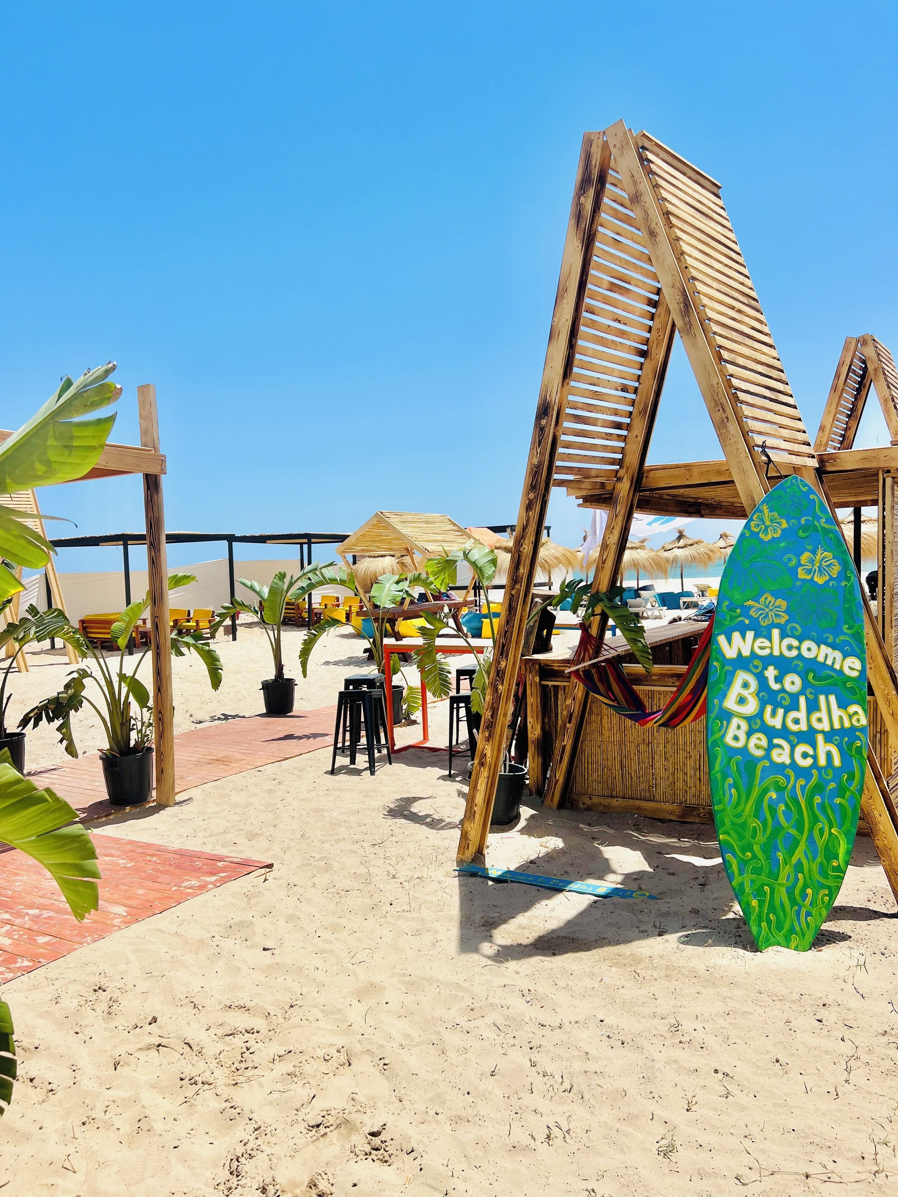 Une zone de plage avec une structure en bois, une planche de surf sur laquelle on peut lire « Bienvenue à Buddha Beach  », et des plantes en pot.