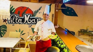 A chef holding plates of food in a restaurant named "Villa Mar" with a wooden ceiling and a sign that reads "VILLAMAR". - Hôtel Villamar (Hammamet)