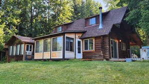 A rustic log cabin with large windows and a chimney, surrounded by a grassy yard and dense trees. - Rustic 4-bedroom, lakefront, log cabin on Bull Lake! (Troy)