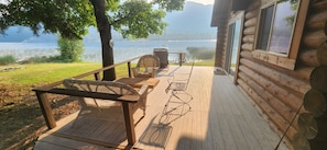 A wooden deck with wicker furniture, a grill, and a view of a lake and mountains. - Rustic 4-bedroom, lakefront, log cabin on Bull Lake! (Troy)