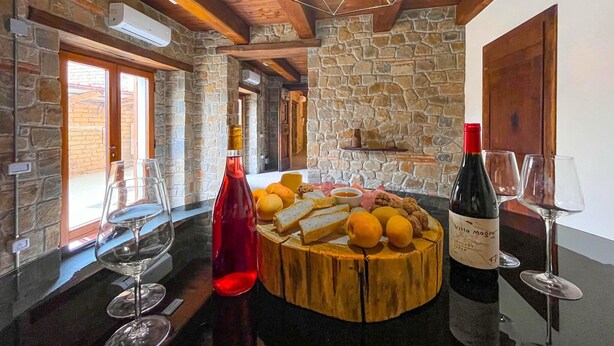 A rustic interior with stone walls, wooden beams, and a round wooden table with food and wine.