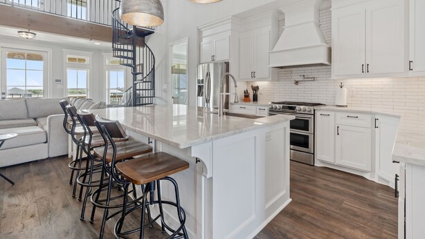 A spacious kitchen with white cabinetry, a central island with bar stools, a stainless steel refrigerator, and a staircase leading to an upper level.