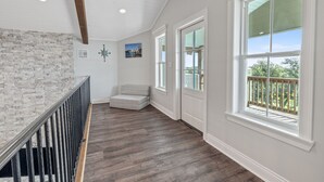 A spacious hallway with a wooden floor, white walls, and large windows offering a view of the outdoors.