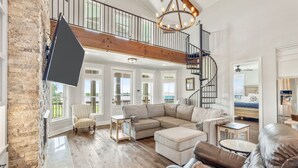 A spacious living room with a large sectional sofa, a leather armchair, and a small table. A wooden staircase leads to an upper level.