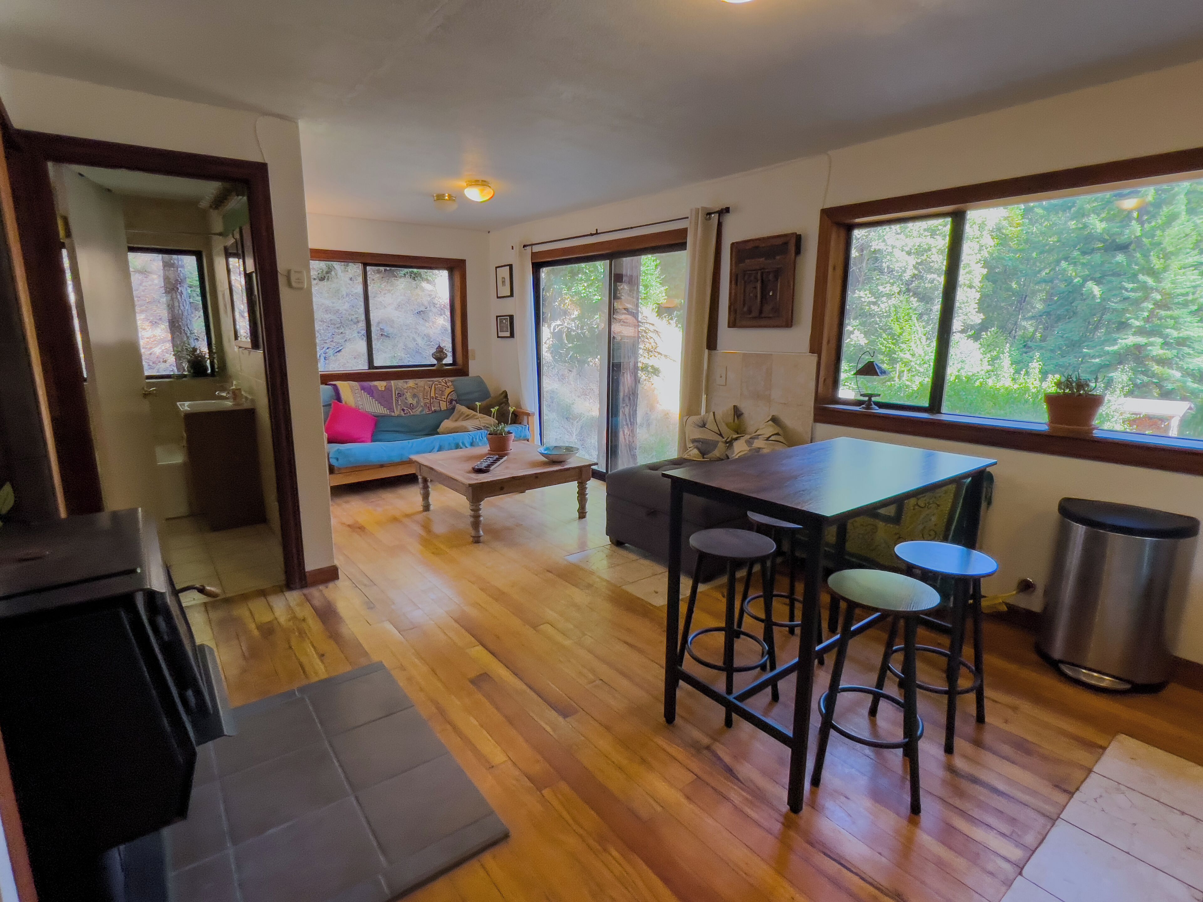 Un salon chaleureux avec un plancher en bois, un canapé, une table basse et une table à manger avec des tabourets. De grandes fenêtres laissent entrer la lumière naturelle et offrent une vue sur la verdure environnante.