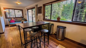 Una cocina con una mesa de madera, taburetes de bar, un cubo de basura y ventanas amplias con vistas al bosque.