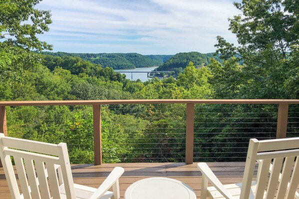 Um terraço com duas cadeiras brancas e uma pequena mesa, com vista para um rio e colinas arborizadas.