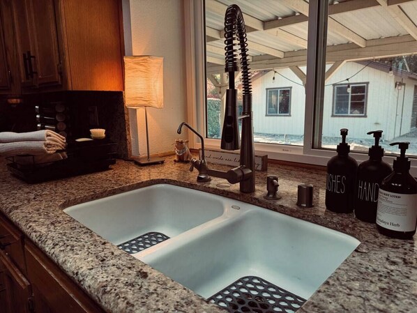 A kitchen with a double sink, granite countertop, and a window with a view of a house.