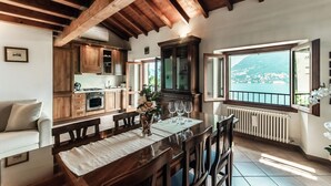 A dining area with a wooden table and chairs, a kitchen with wooden cabinets, and a large window with a view of a body of water and hills. - Lake Frame - MyHomeinComo (Blevio)