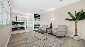A modern living room with a round rug, a coffee table, a grey armchair, a sofa, a built-in bookshelf with greenery, and a potted plant.