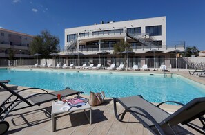 Une piscine avec des chaises longues, une table avec une boisson et un sac, et un bâtiment doté d’un escalier et de balcons.