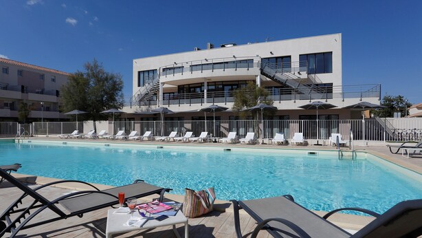 Zona de piscina con sillones de descanso y sombrillas, un edificio moderno con una escalera, y un cielo azul despejado.