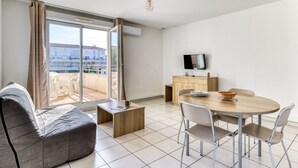 A modern living room with a sofa, a dining table with chairs, a TV on a wooden cabinet, and a balcony with a view of buildings.