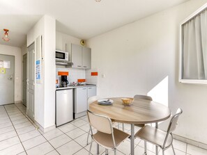 A compact kitchen with a microwave, coffee maker, and a small refrigerator. There is a dining table with chairs and a window with curtains.