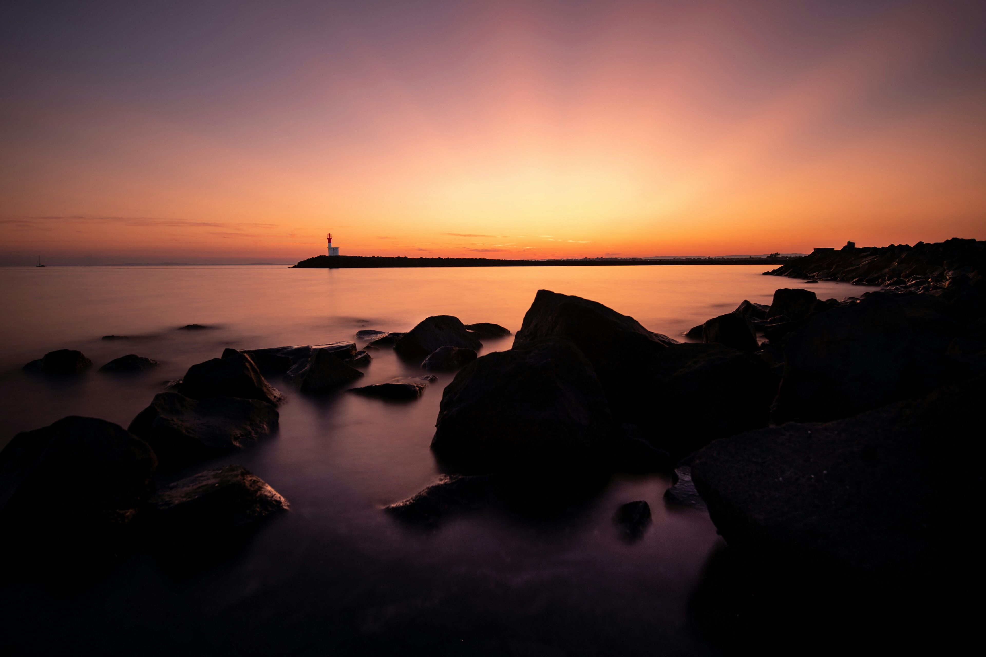 Un coucher de soleil sur la côte, avec la silhouette d’un phare, une mer calme et un rivage rocheux.