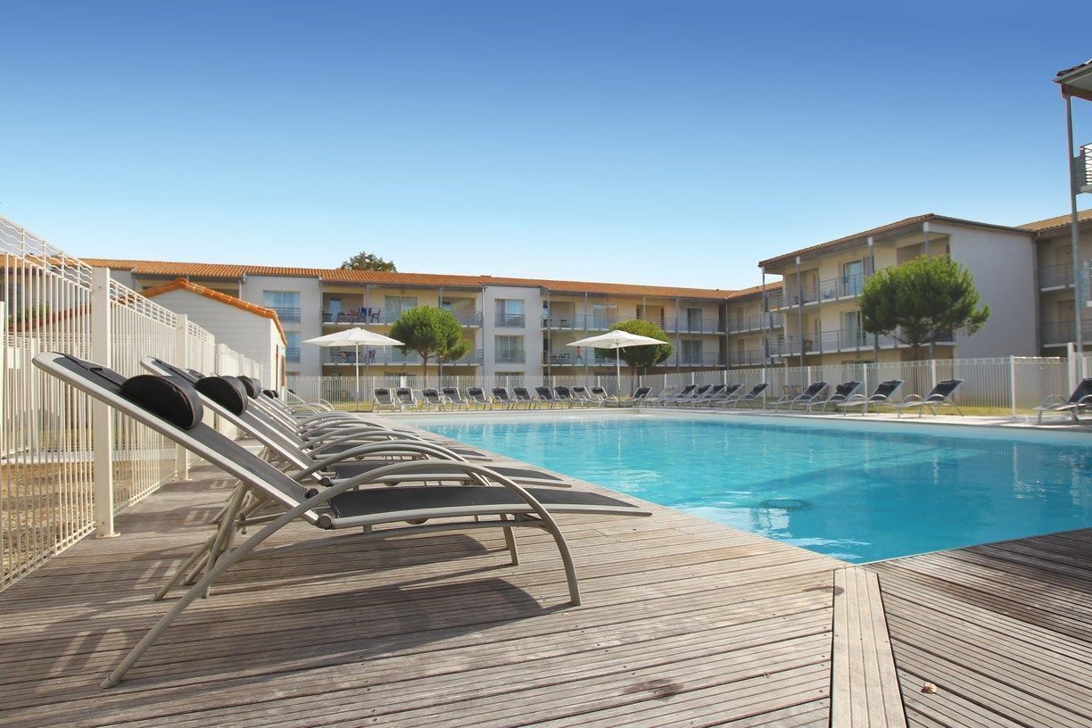 Ein Poolbereich mit Liegestühlen und Sonnenschirmen, eine Holzterrasse und ein Gebäude mit Balkonen im Hintergrund.