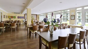 A spacious dining area with wooden tables and chairs, large windows offering a view of the outside, and a variety of framed artworks on the walls.