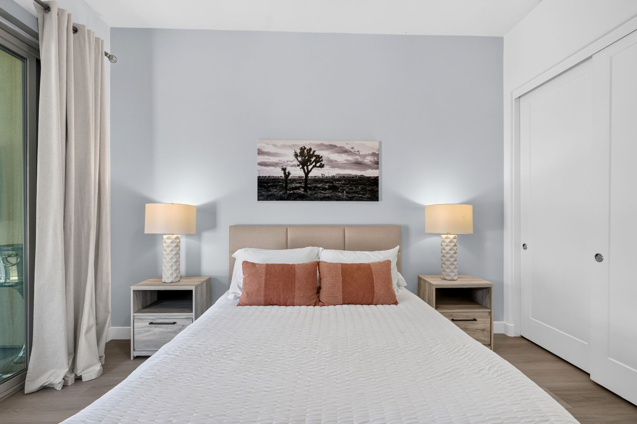 Une chambre à coucher avec un lit, deux tables de chevet avec des lampes, une commode, une fenêtre avec des rideaux et un tableau accroché au mur.