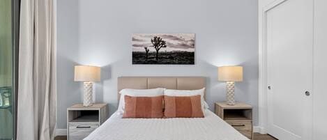 Une chambre à coucher avec un lit, deux tables de chevet avec des lampes, une commode, une fenêtre avec des rideaux et un tableau accroché au mur.