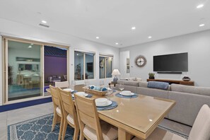 A modern living room with a dining area, a flat-screen TV mounted on the wall, and a large sliding glass door leading to an outdoor space. - El Encanto: EV Charger, Pool, Spa & BBQ (La Quinta)