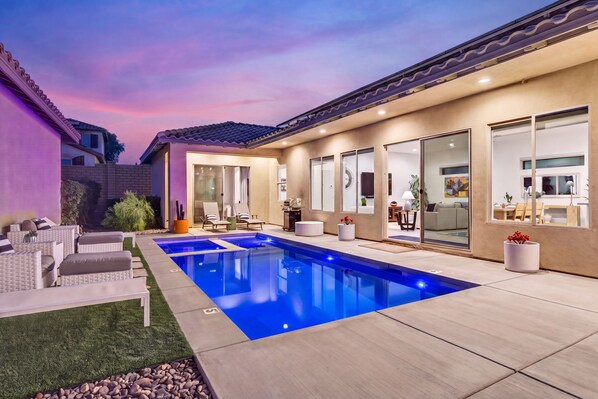 A modern outdoor swimming pool with a patio area, seating, and a view into the house.